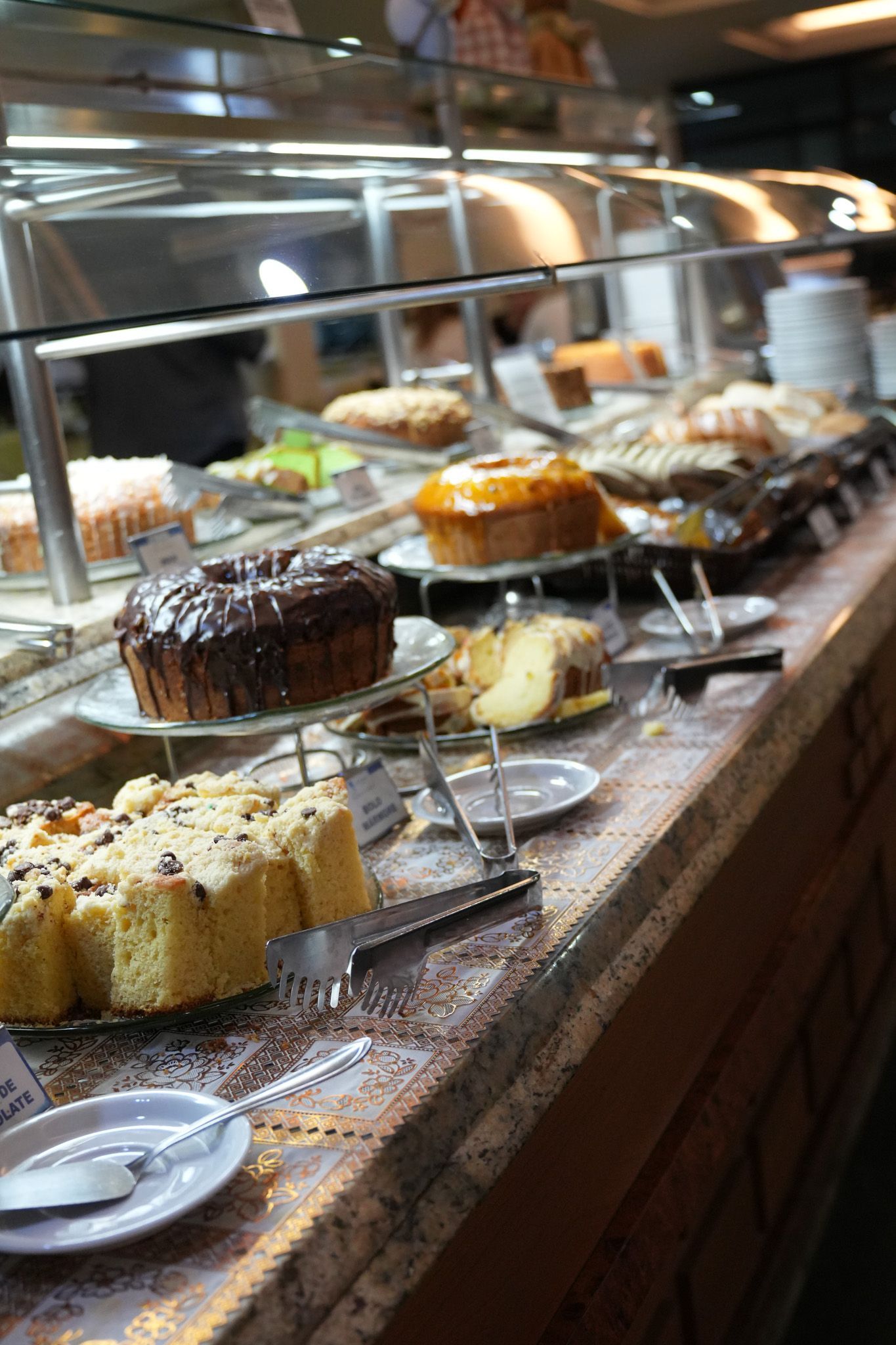 Buffet de sobremesas com bolos e doces dispostos em um balcão de restaurante sob uma vitrine de vidro.