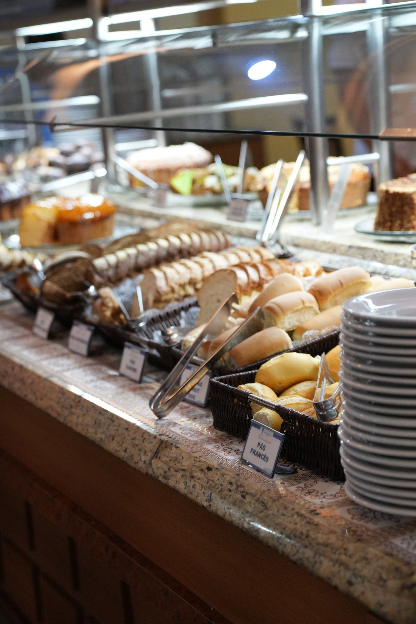 Buffet de padaria com uma variedade de doces em bandejas, pegadores e pratos empilhados sobre um balcão de granito.