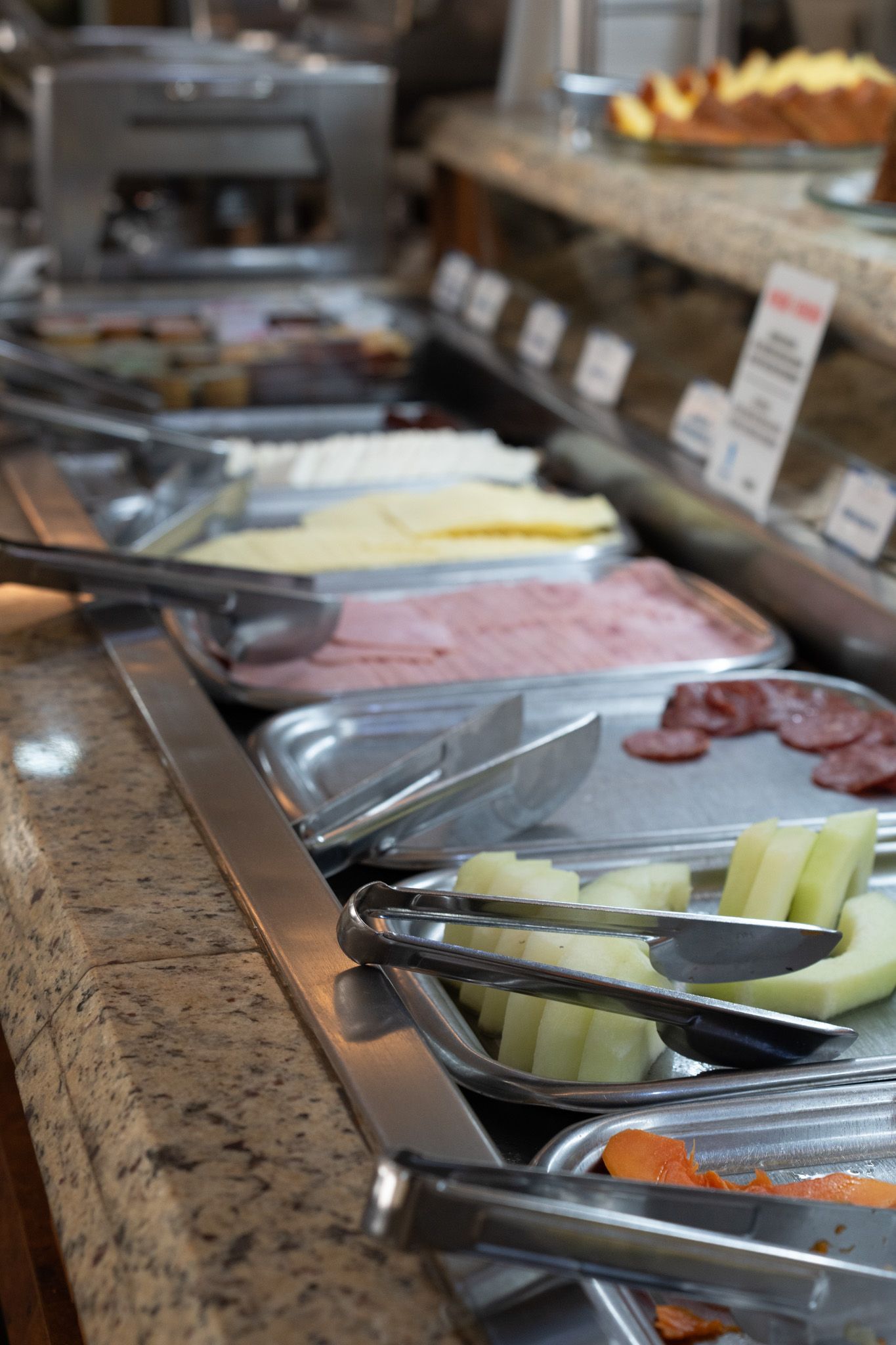 Balcão de buffet com pegadores de metal e bandejas de frutas fatiadas, queijos e frios sobre uma bancada de granito.
