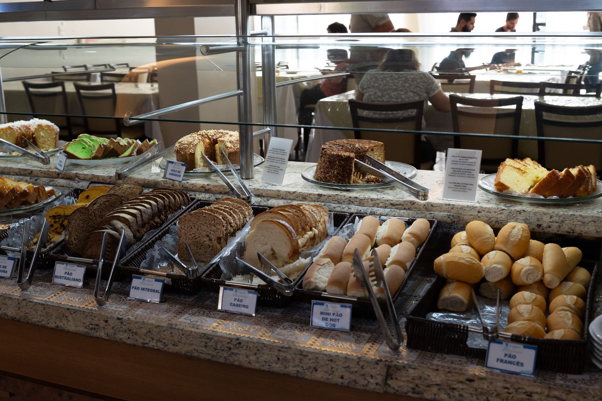 Vitrine de padaria com uma variedade de doces e bolos em bandejas, com mesas de café ao fundo.