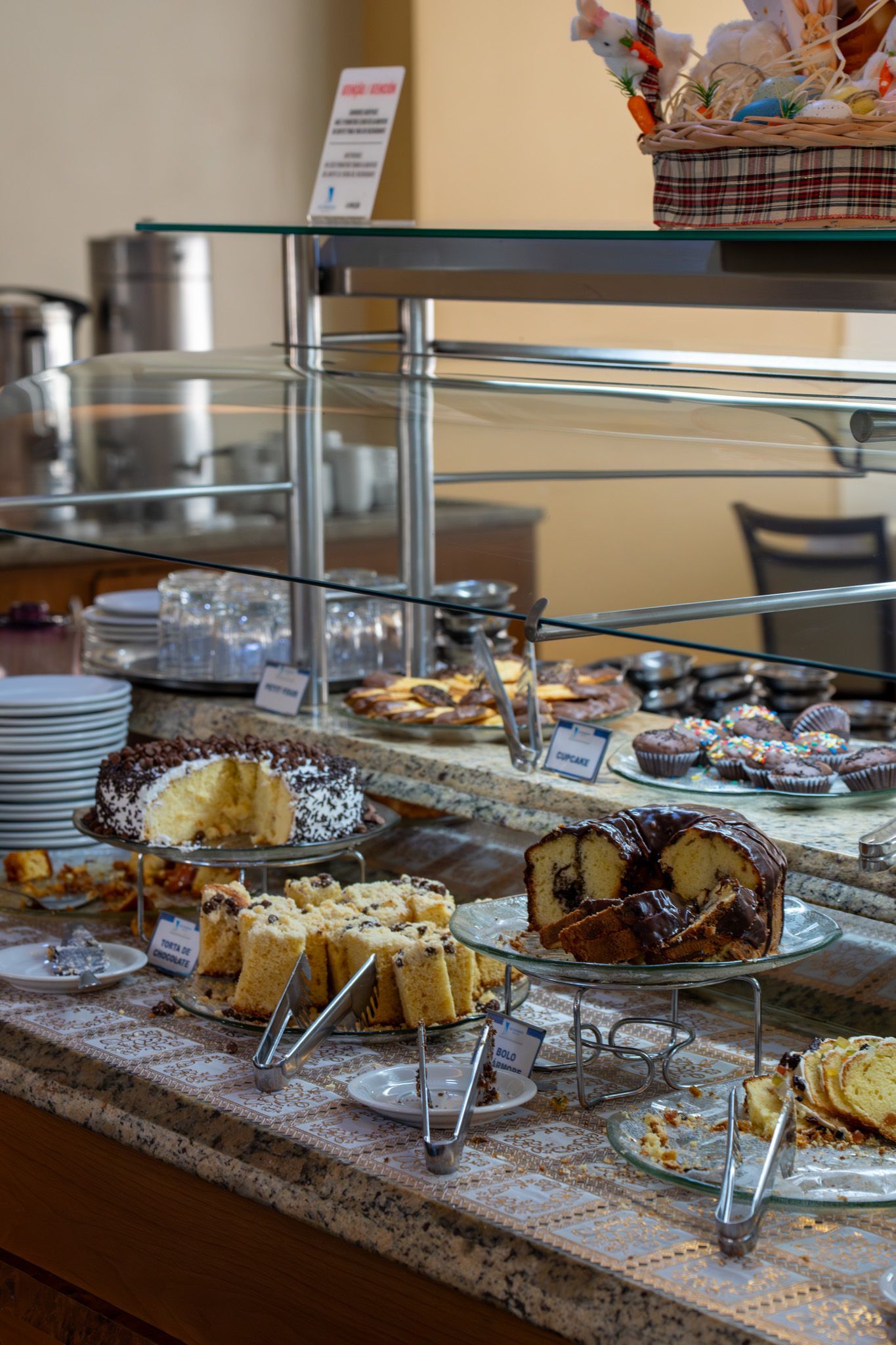 Doces expostos em um balcão de exposição em um café, com bandejas de sobremesas e uma vitrine de vidro.