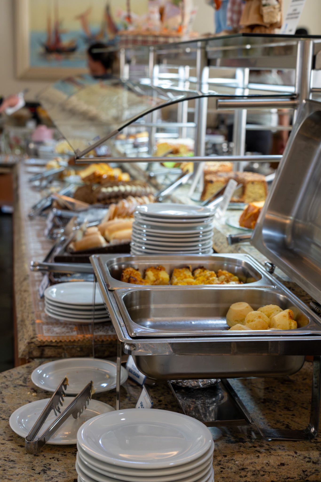 Mesa de buffet com bandejas de aço inoxidável contendo alimentos, pratos e utensílios para servir, sobre uma bancada de granito.