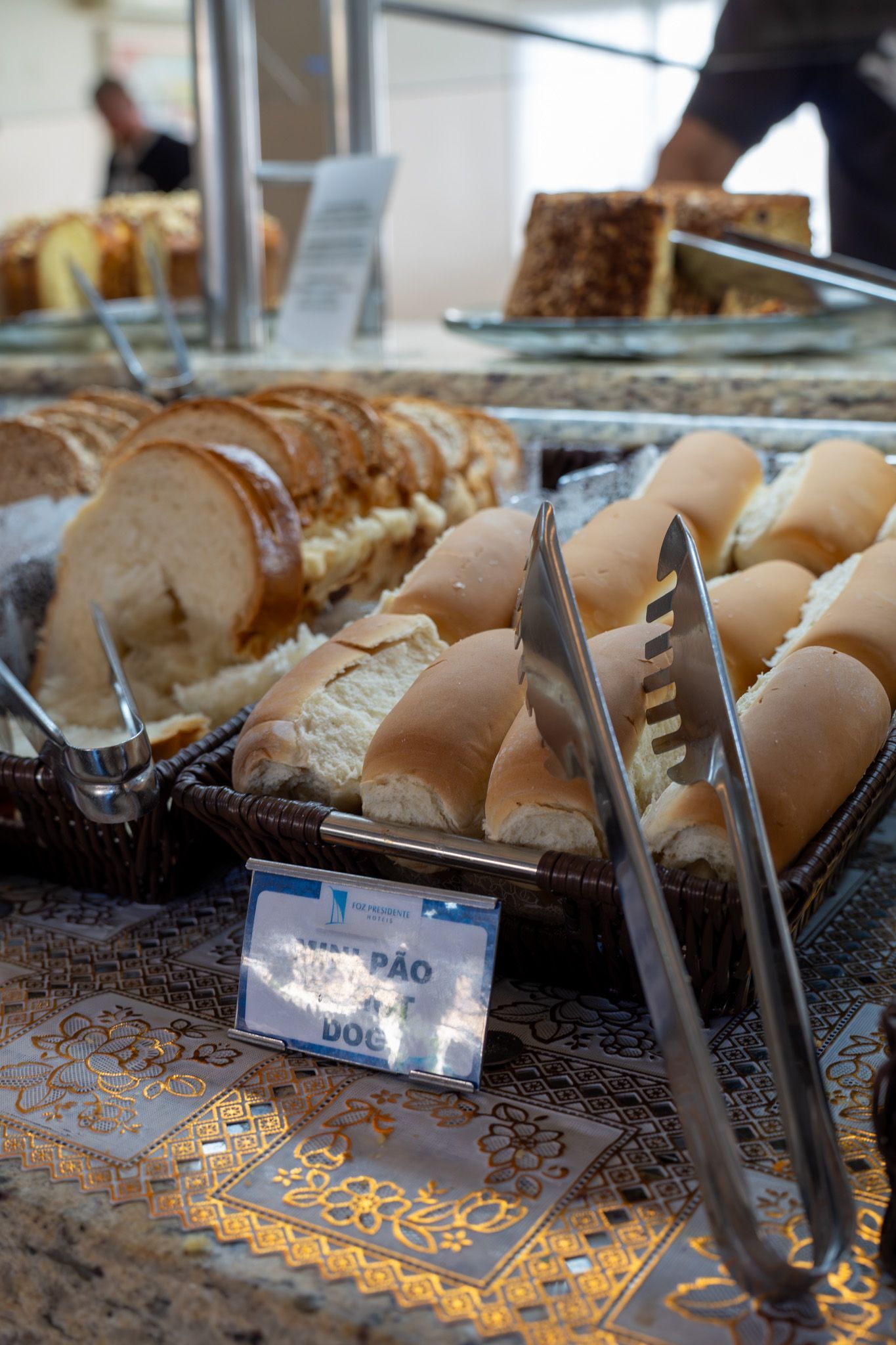 Buffet de padaria com pão fatiado, pãezinhos e pegadores sobre uma mesa com toalha estampada.