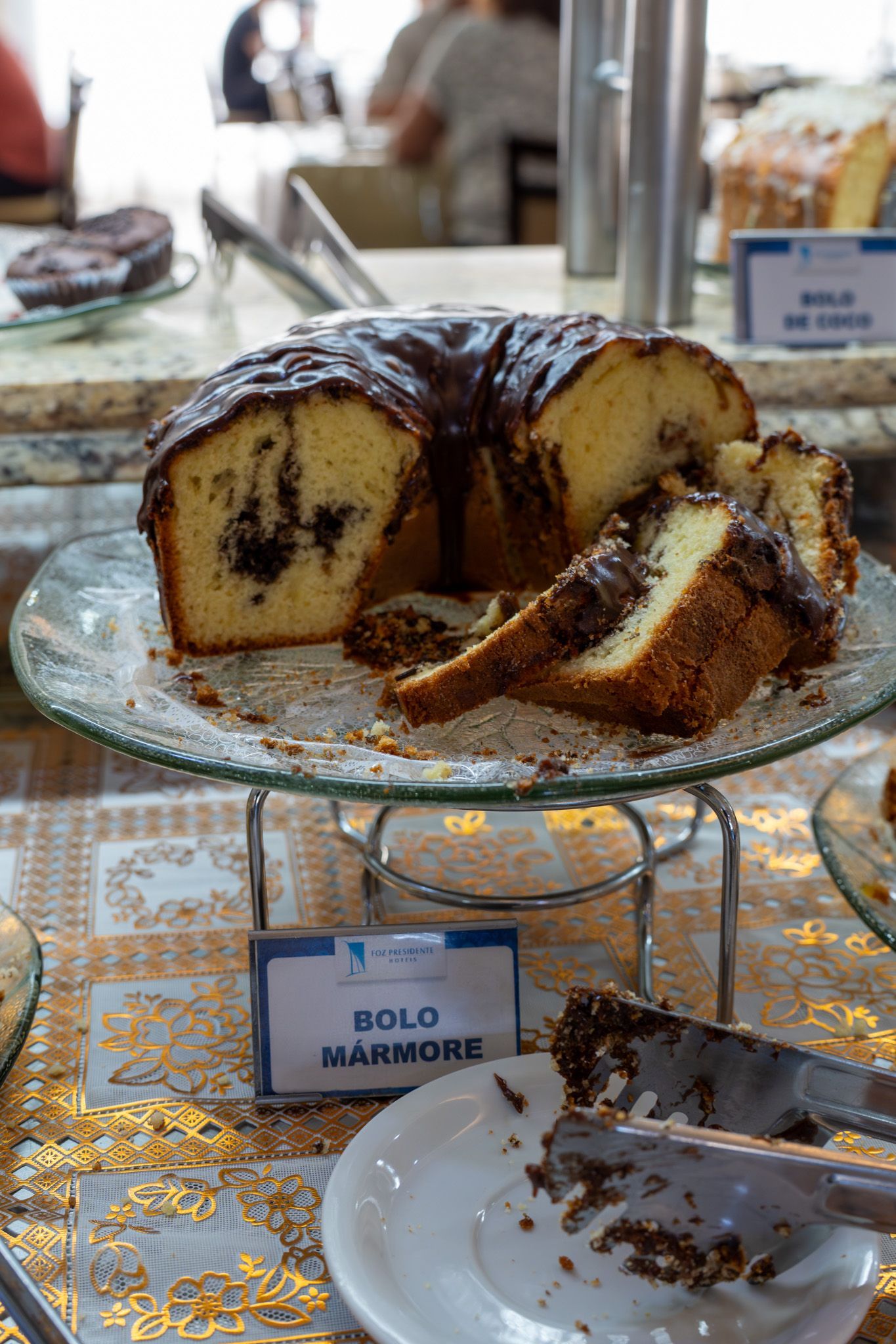 Fatias de bolo de chocolate em um suporte de vidro com uma toalha de mesa salpicada de dourado e pratos de sobremesa abaixo.