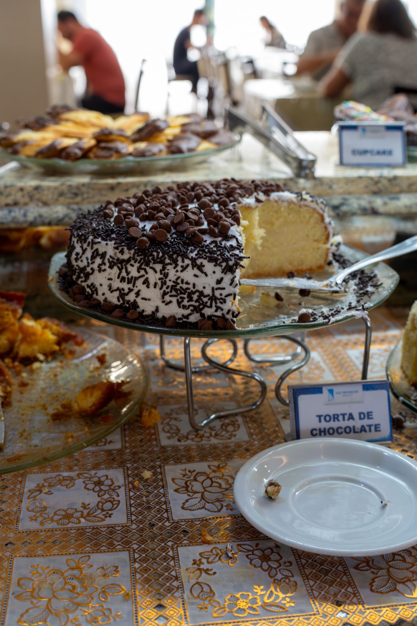 Bolo de chocolate em uma mesa de buffet com etiqueta e uma fatia faltando, em uma sala de jantar.