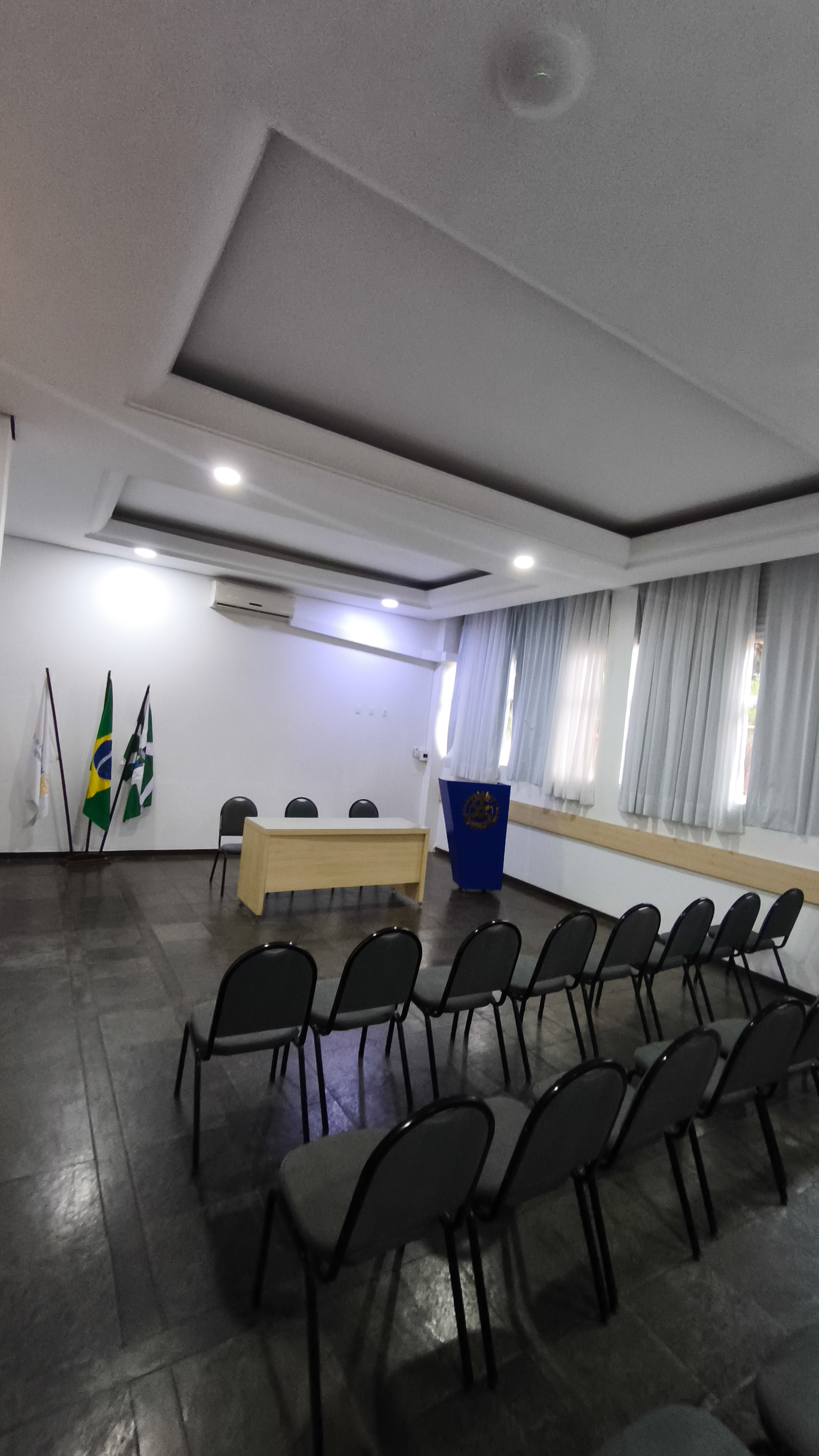 Sala de conferências vazia com fileiras de cadeiras pretas, um pódio, bandeiras e uma mesa na frente.