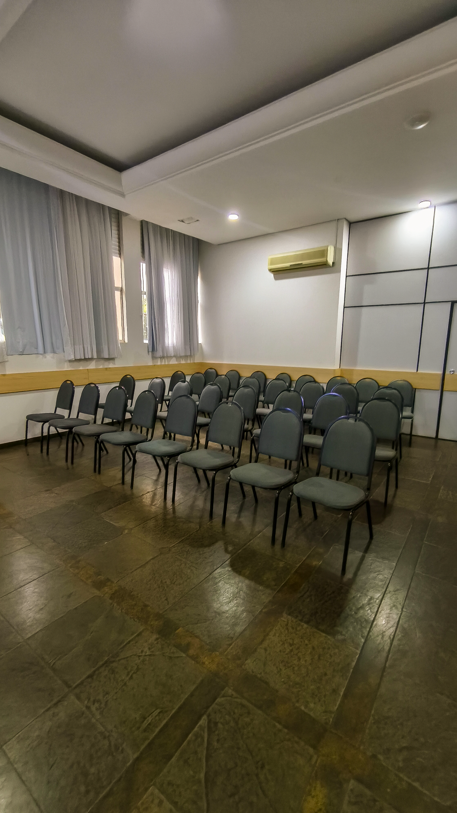 Sala de conferências vazia com fileiras de cadeiras verdes, piso de azulejo, janelas e ar-condicionado de parede.