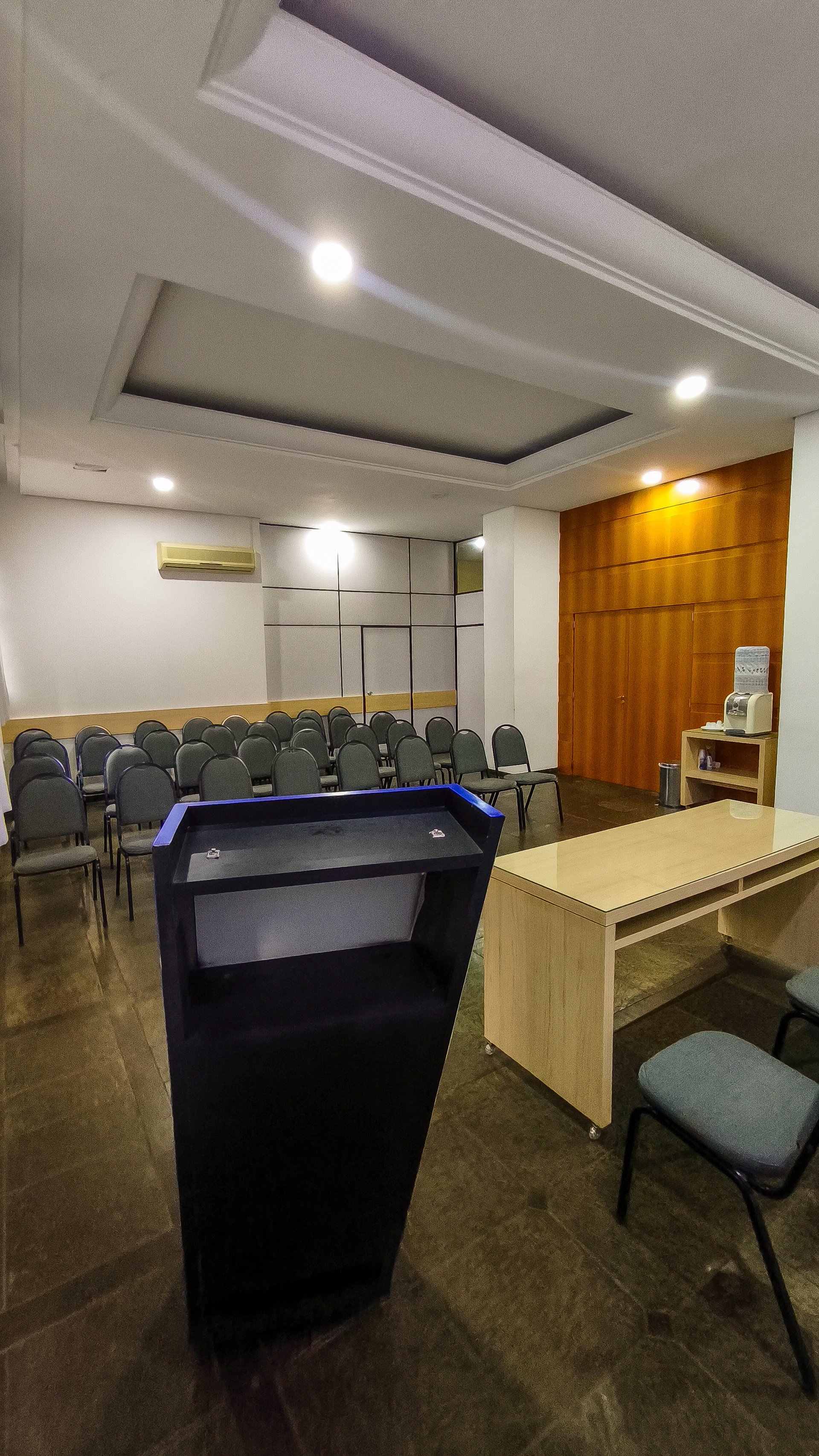 Tribunal vazio com banco de madeira, pódio, fileiras de cadeiras e paredes brancas sob iluminação embutida.