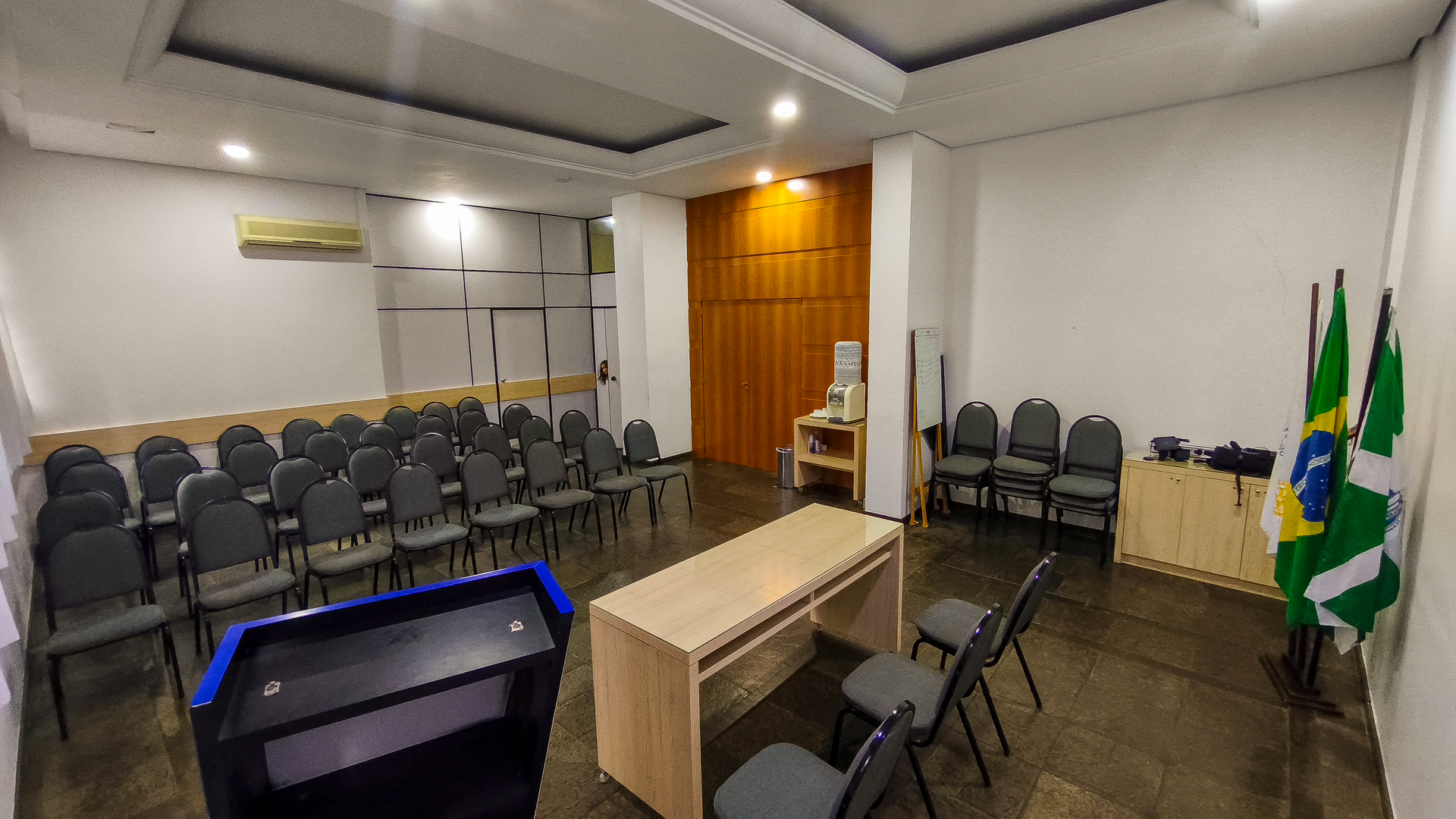 Sala de conferências vazia com fileiras de cadeiras, uma mesa, um pódio e bandeiras perto da parede frontal.
