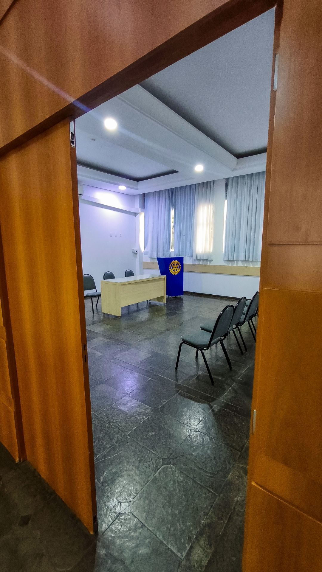 Vista através de uma porta para uma pequena sala de reuniões com cadeiras, um pódio, paredes brancas e janelas com cortinas.