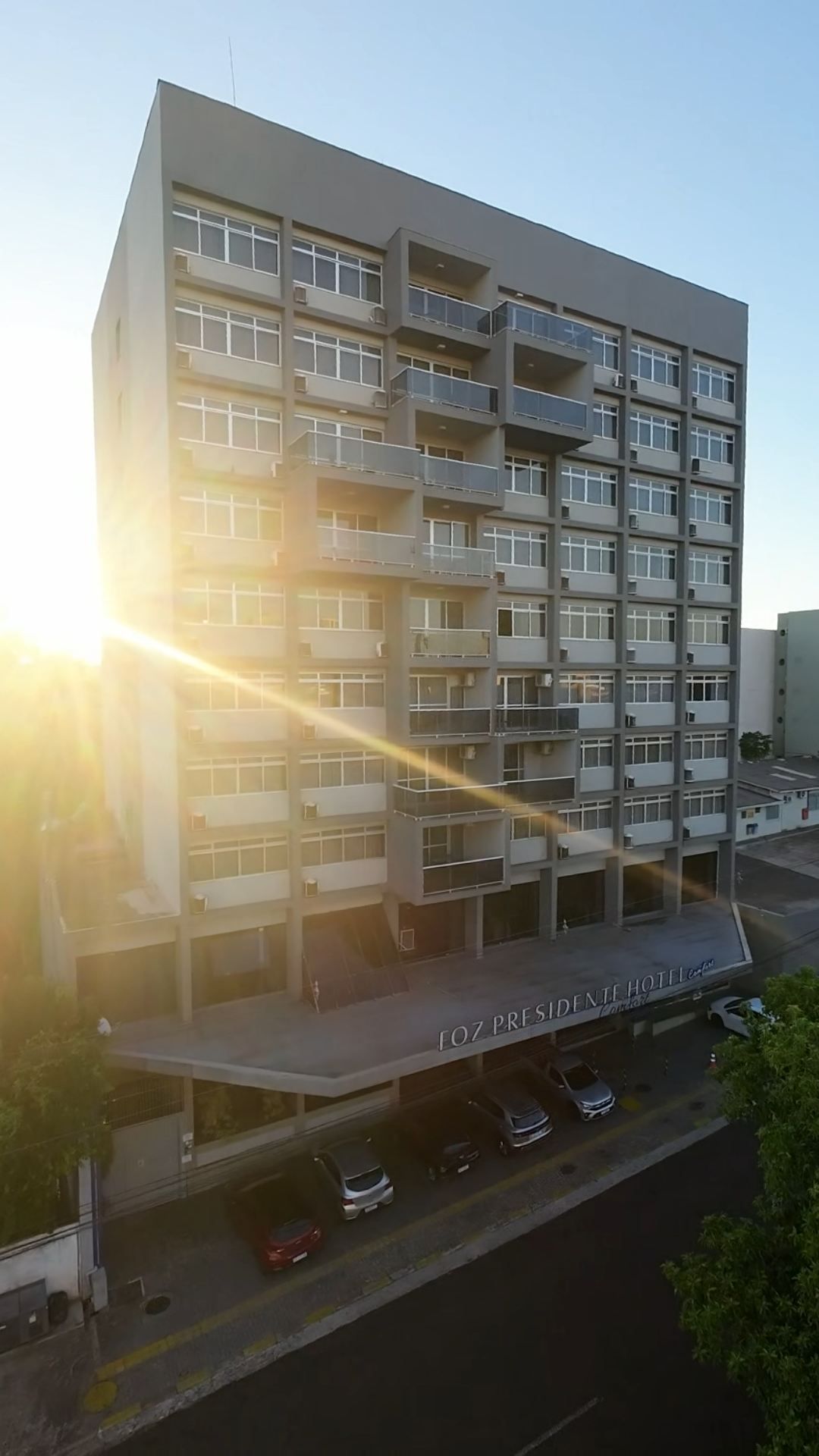 Prédio residencial moderno de altura média ao pôr do sol, com forte reflexo do sol e carros estacionados embaixo.