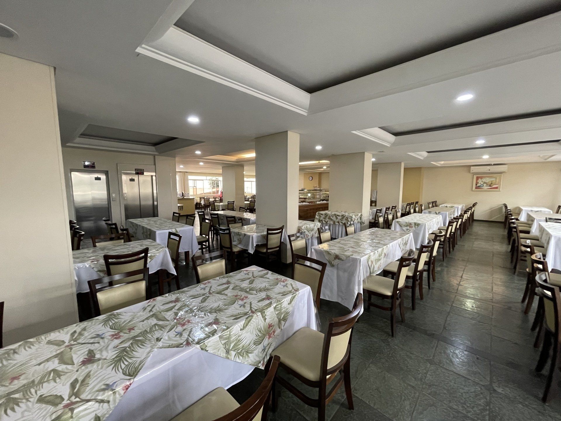 Sala de jantar vazia de um restaurante com mesas cuidadosamente arrumadas, luminárias brancas no teto e paredes cor creme.