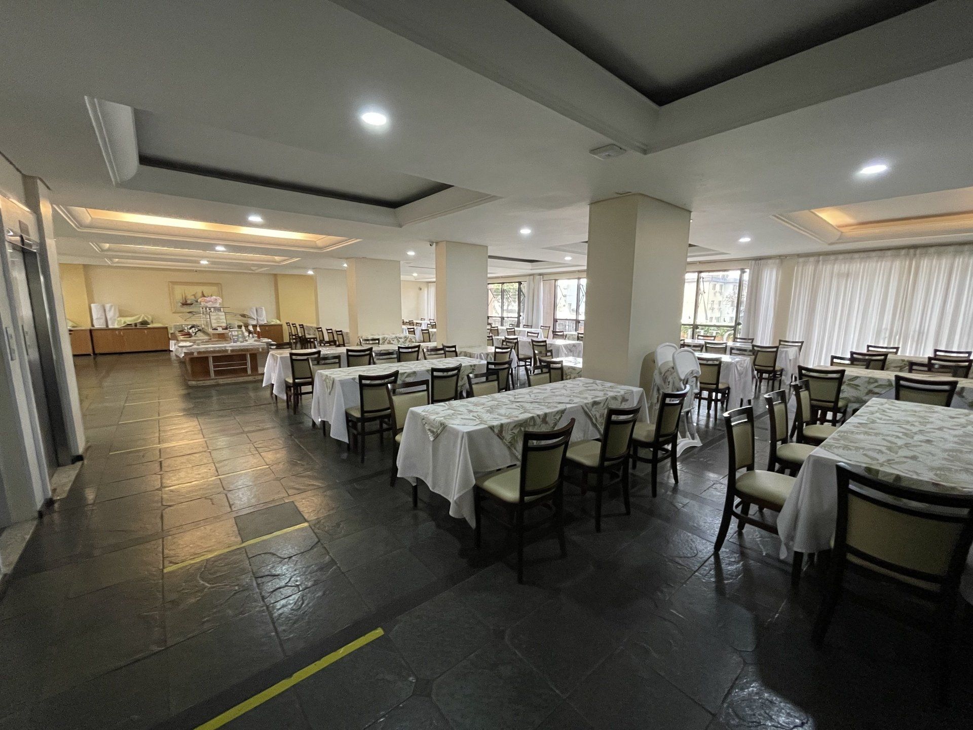 Sala de jantar de restaurante iluminada, com mesas e cadeiras cuidadosamente arrumadas sob luzes embutidas no teto.