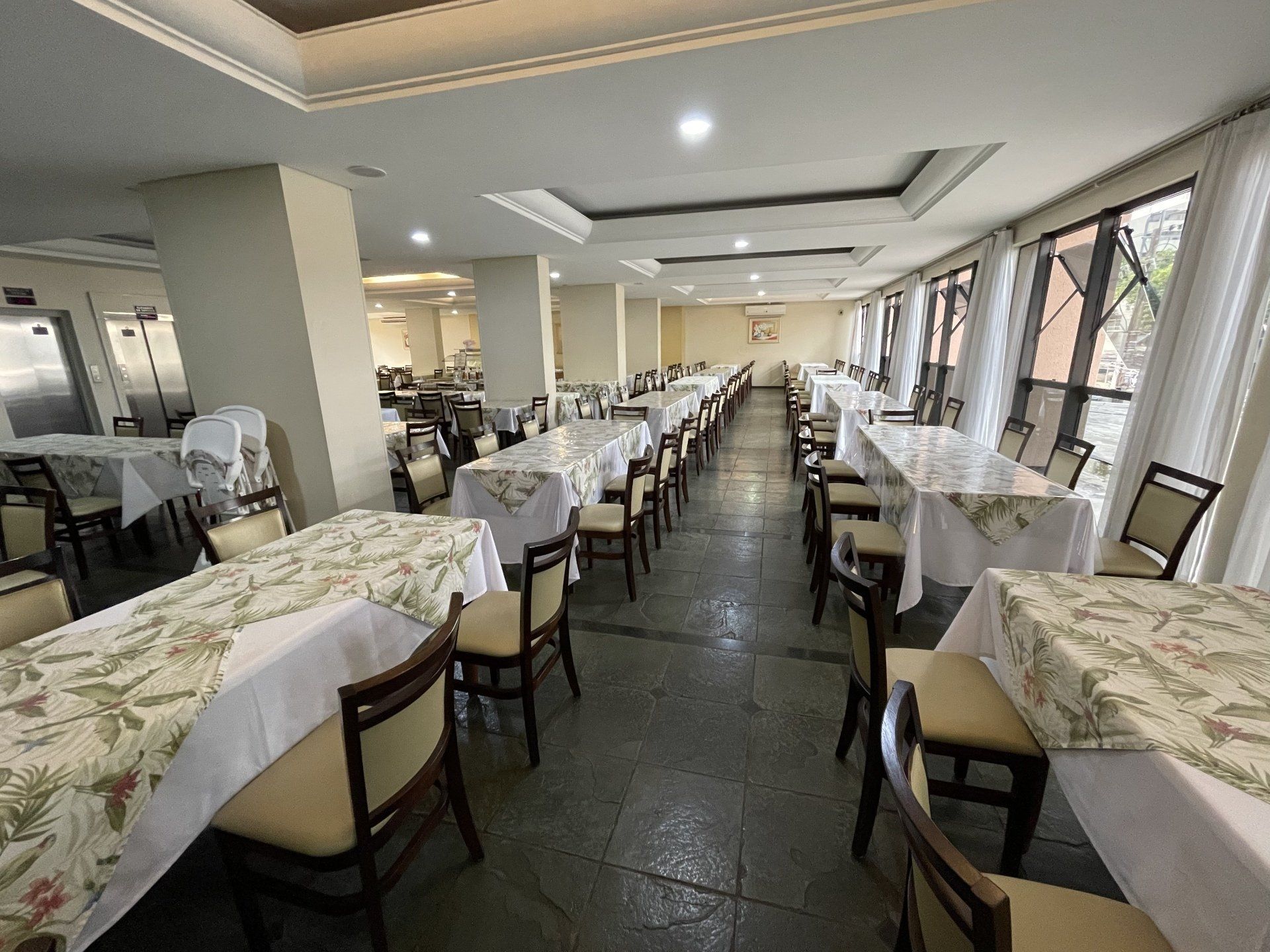 Sala de jantar de restaurante iluminada, com fileiras de mesas postas, toalhas de mesa brancas e cadeiras ao longo de grandes janelas.