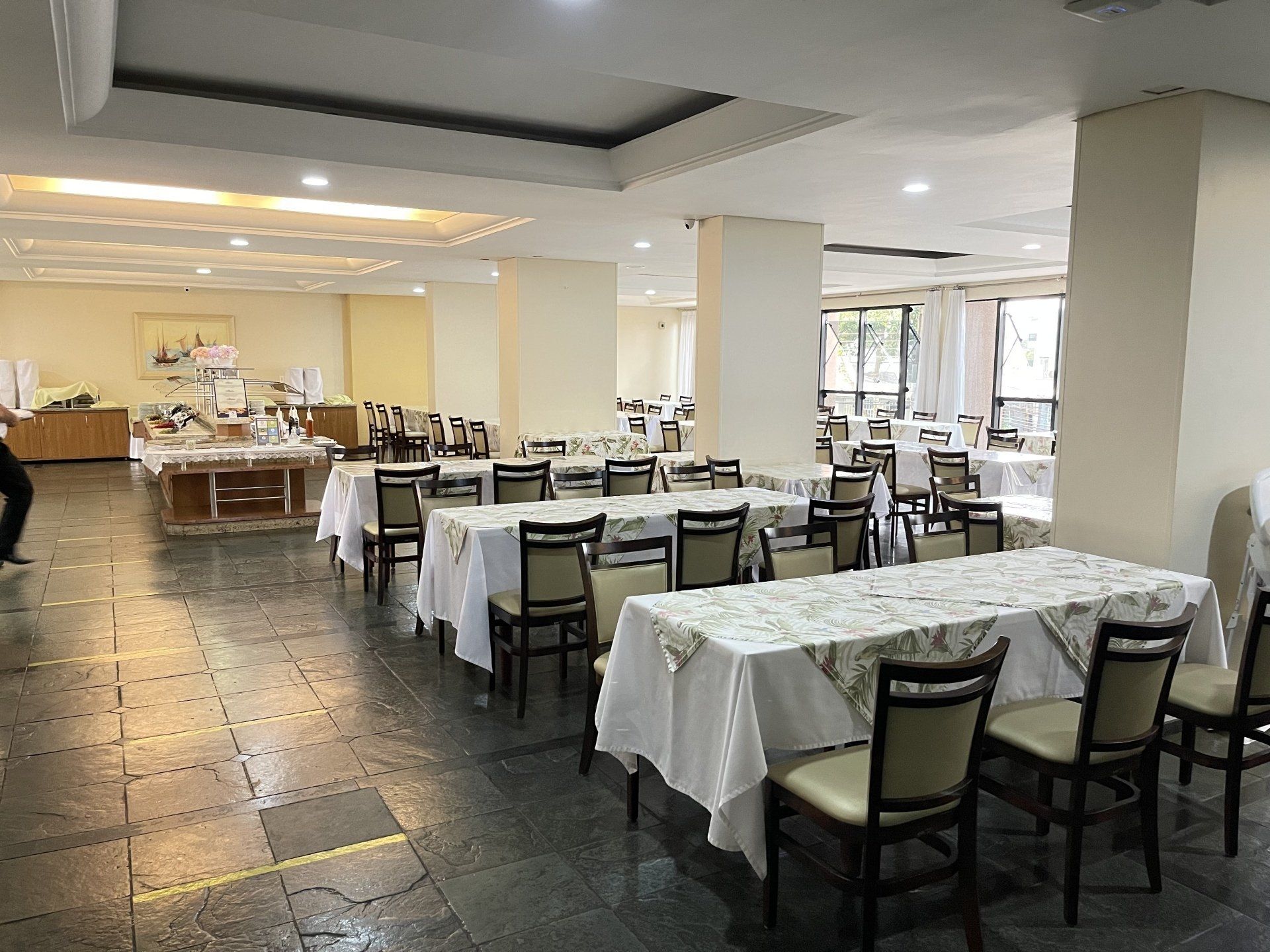 Salão de banquetes vazio com toalhas de mesa brancas, cadeiras pretas e mesas de bufê em uma sala de jantar iluminada.