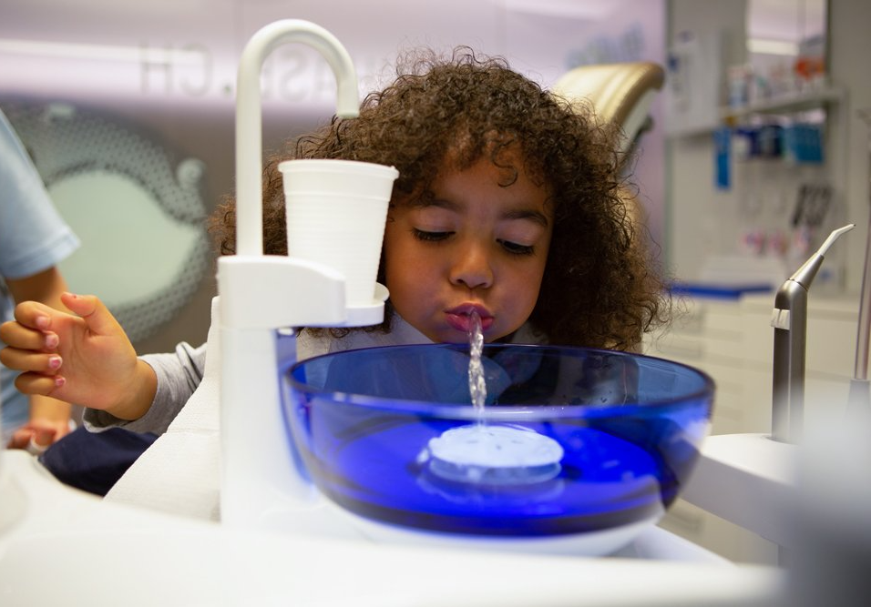 A little girl is drinking water from a faucet in a dental office.