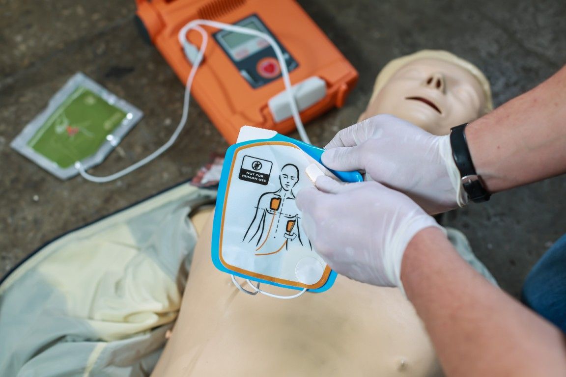 Person applying AED pads to a mannequin's chest, orange AED device in the background.