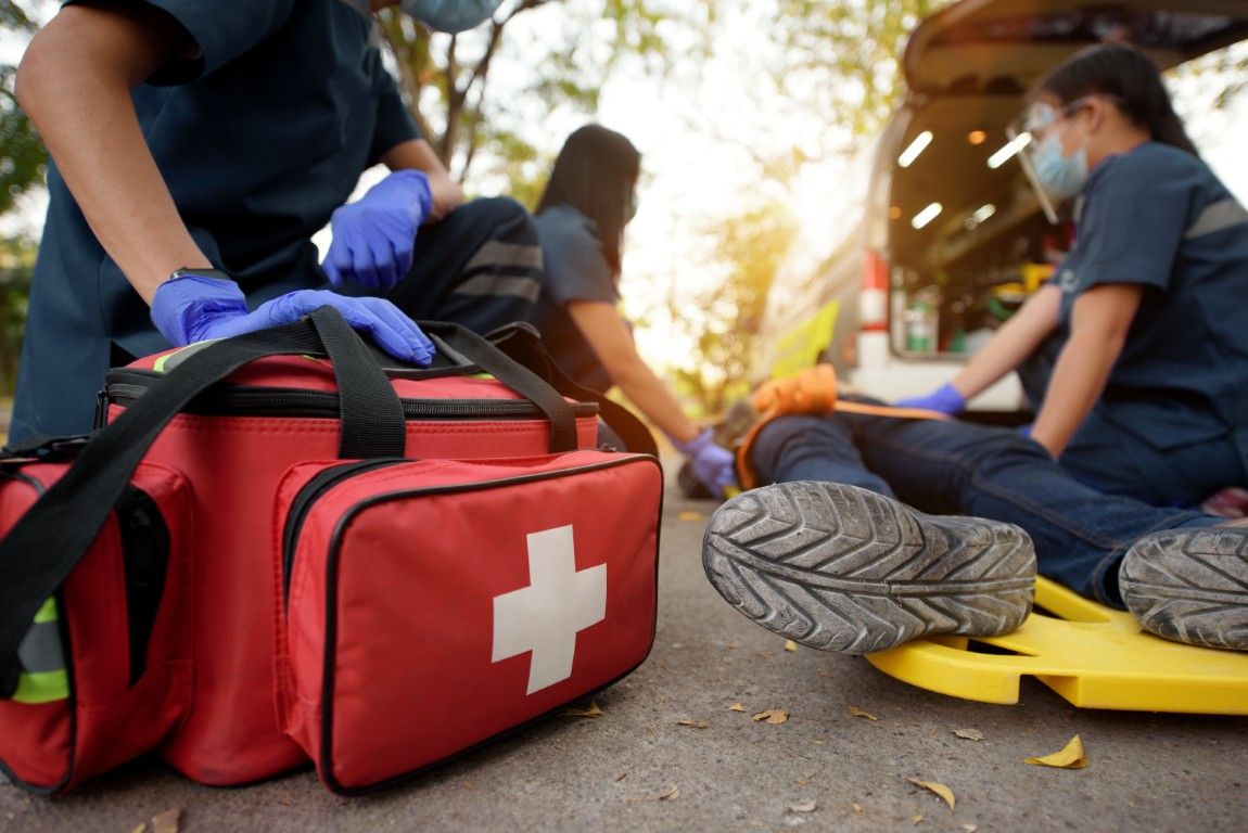 Emergency medical personnel attending to a patient on a stretcher near an ambulance, red medical bag in the foreground.