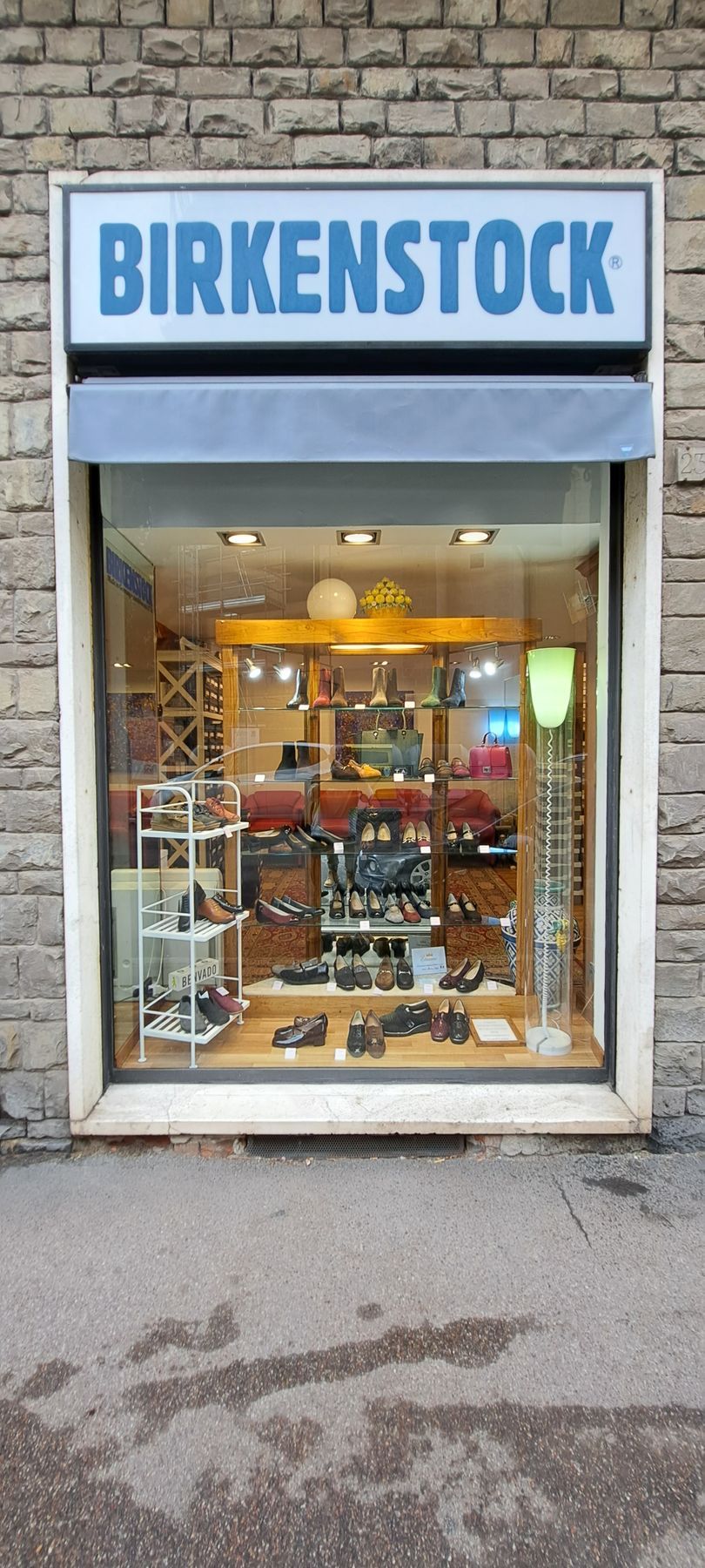 Facciata di un negozio Birkenstock con esposizione di scarpe. Insegna blu sopra il negozio. Tenda grigia e muro di mattoni.