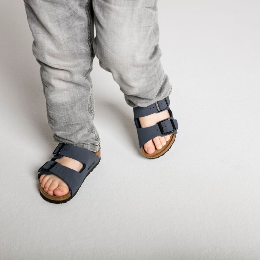 Bambino che indossa jeans grigi e sandali blu, in piedi su una superficie bianca.