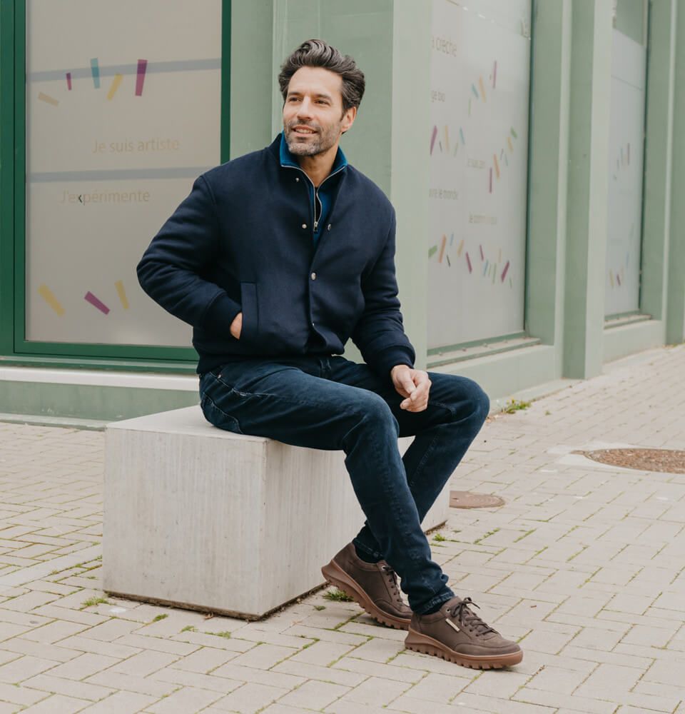 Un uomo con giacca blu, jeans e scarpe marroni è seduto su un blocco di cemento davanti a un negozio e guarda di lato.