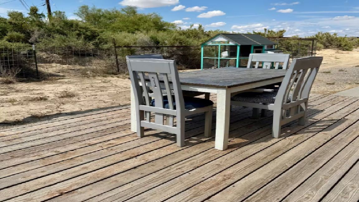 Outdoor dining table with four gray chairs on wooden deck; a chicken coop and shrubs in the background.