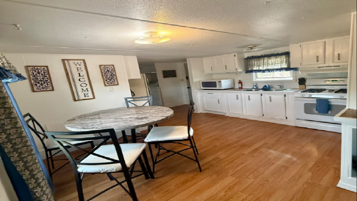 Interior of a mobile home: kitchen with white cabinets, table with chairs, and light brown wood floors.