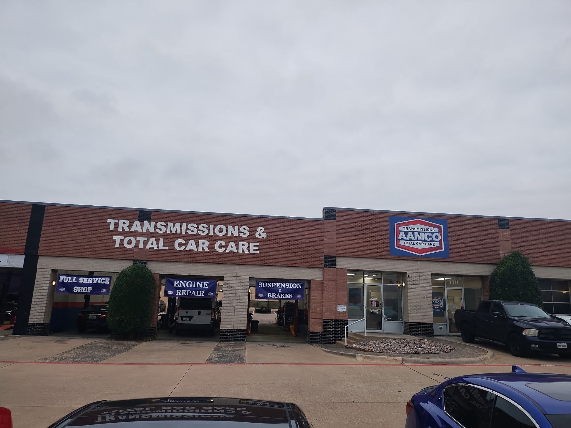 AAMCO Carrollton, TX- Transmission and auto repair shop storefront with brick facade, “Transmissions & Total Car Care” sign, and parked cars under cloudy sky