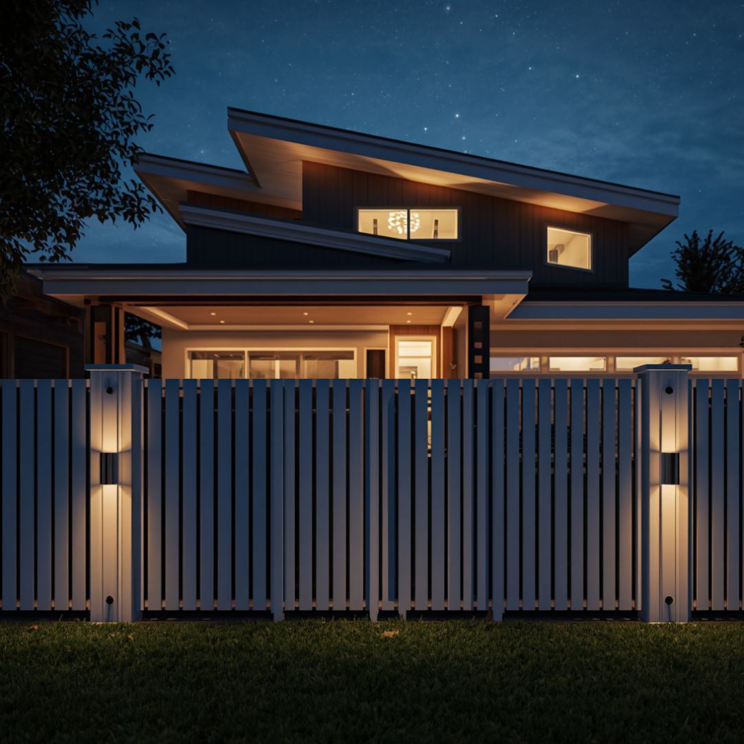 Modern house at night behind a white slat fence, with warm interior lights and illuminated gate posts in woodbury mn
