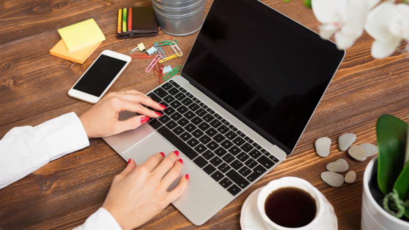 Hands typing on laptop keyboard at a wooden desk, smartphone, coffee, and plant nearby.