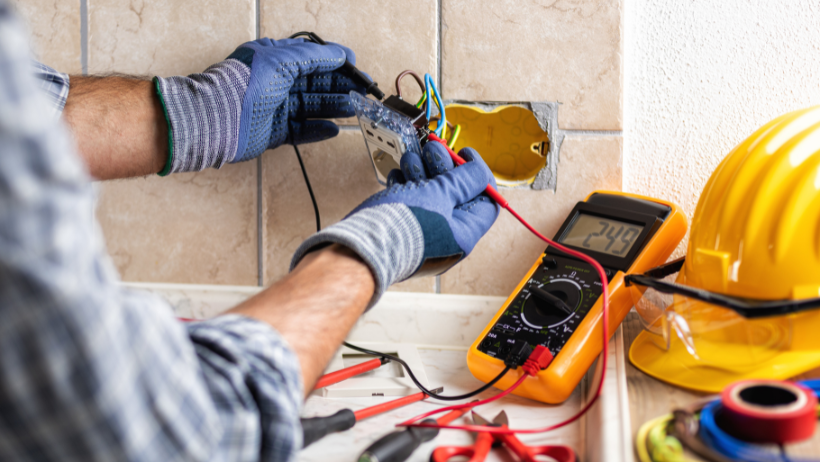 Electrician using a multimeter on electrical wires inside a wall outlet box. Yellow hard hat and tools nearby.