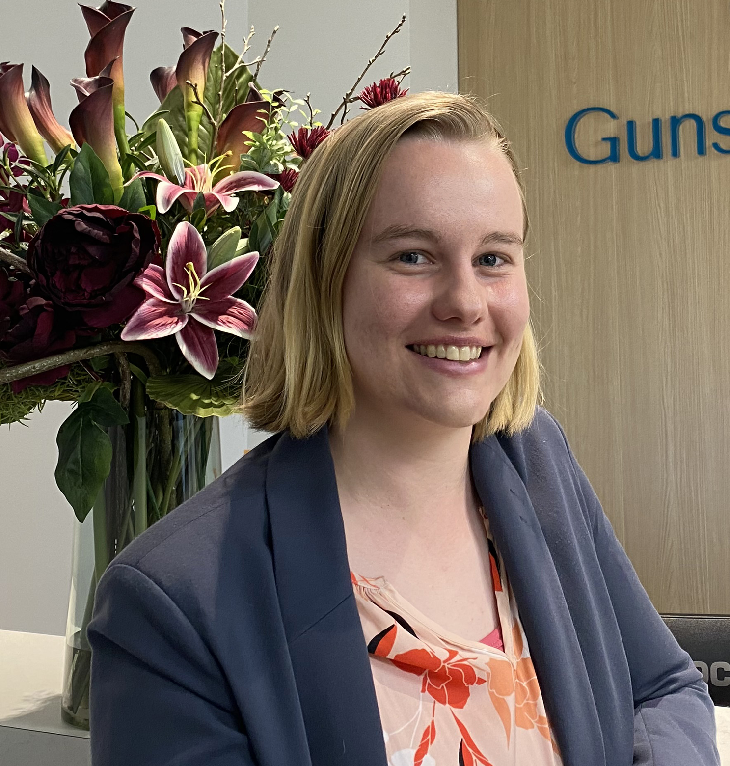 Woman with short blonde hair smiles, wearing a blazer. Flowers in a vase are behind her. 