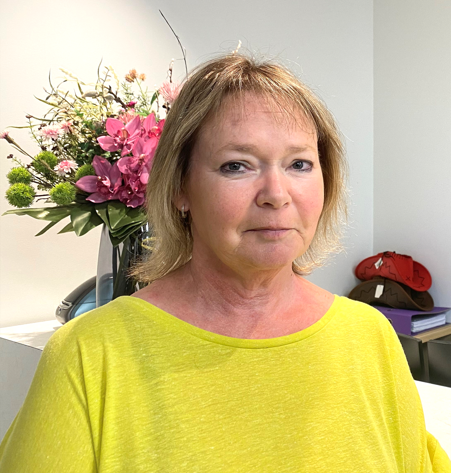 Woman in yellow shirt, looking at the camera. Floral arrangement and desk visible in the background.