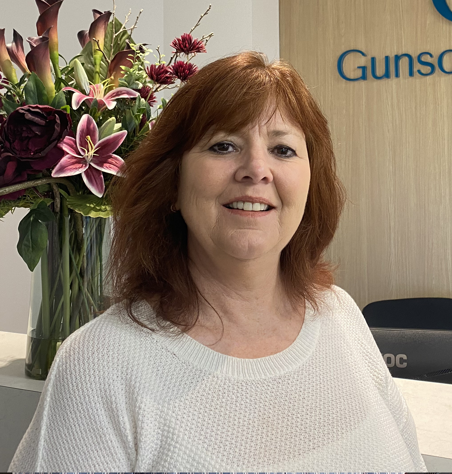 Woman with red hair smiling, standing in front of flowers and a sign that says 