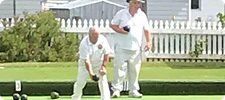 Two men in white uniforms playing lawn bowls on a green lawn with a white fence.