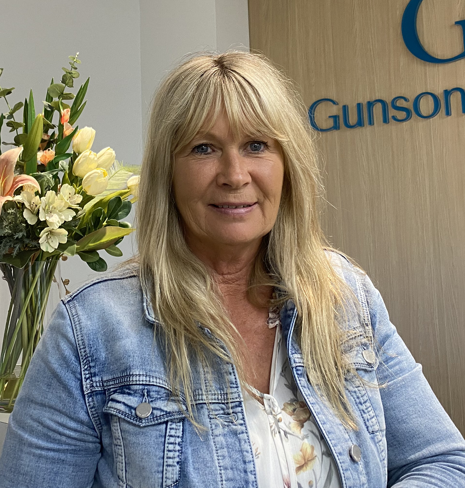 Woman in denim jacket smiles, in an office with flowers. 