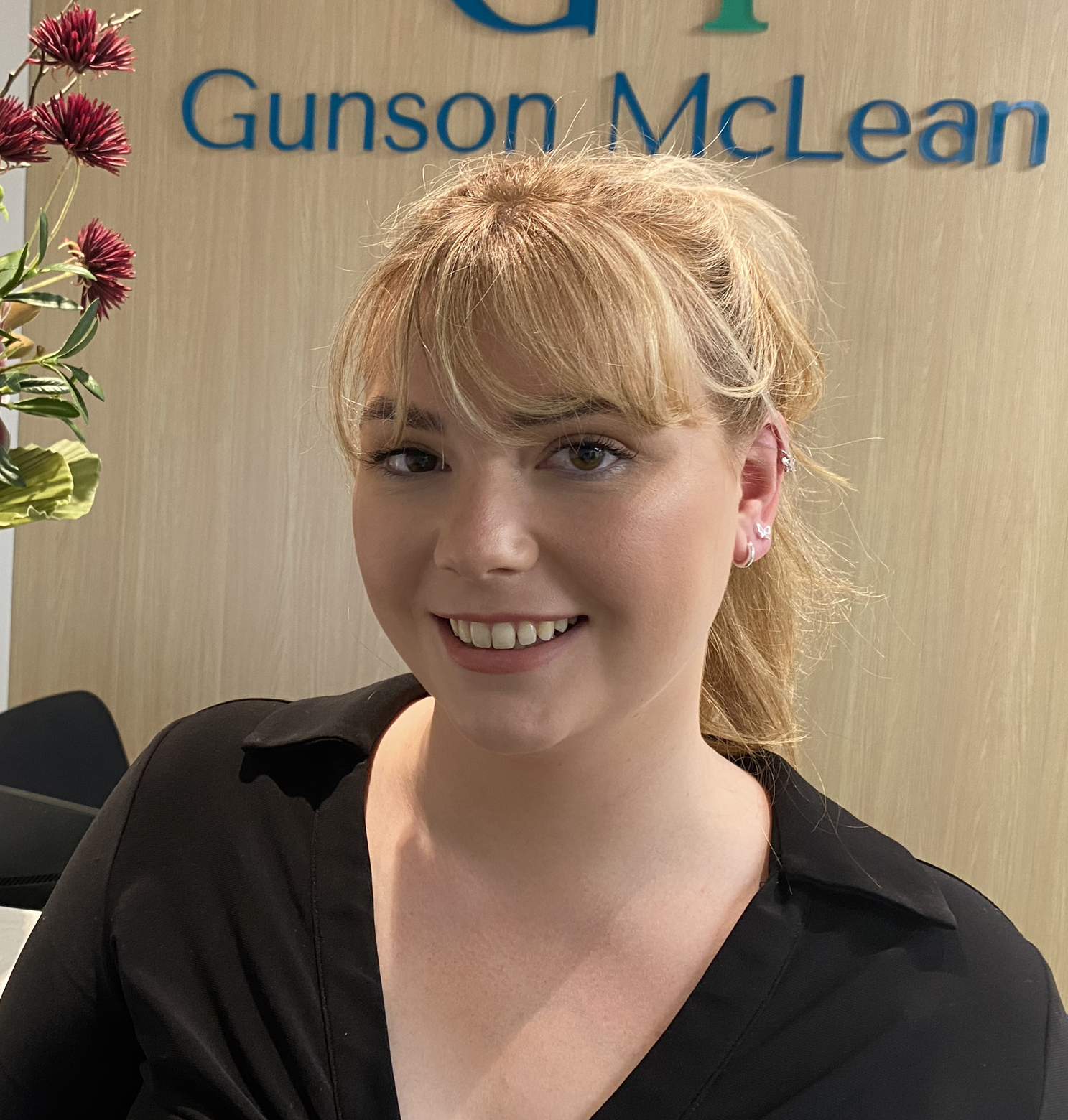 Woman in black shirt smiles at the camera, in front of Gunson McLean sign and flowers.