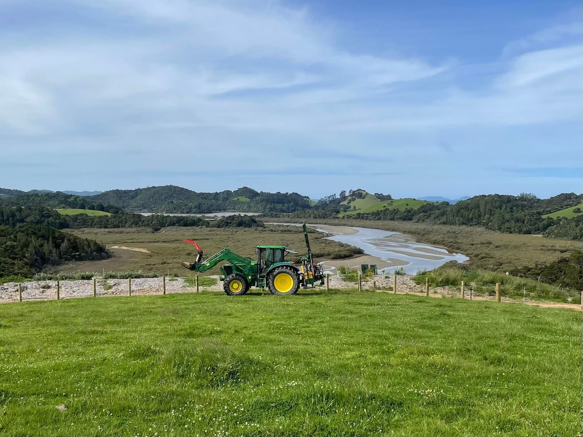 Green tractor on a grassy hill overlooking a river valley under a partly cloudy sky.