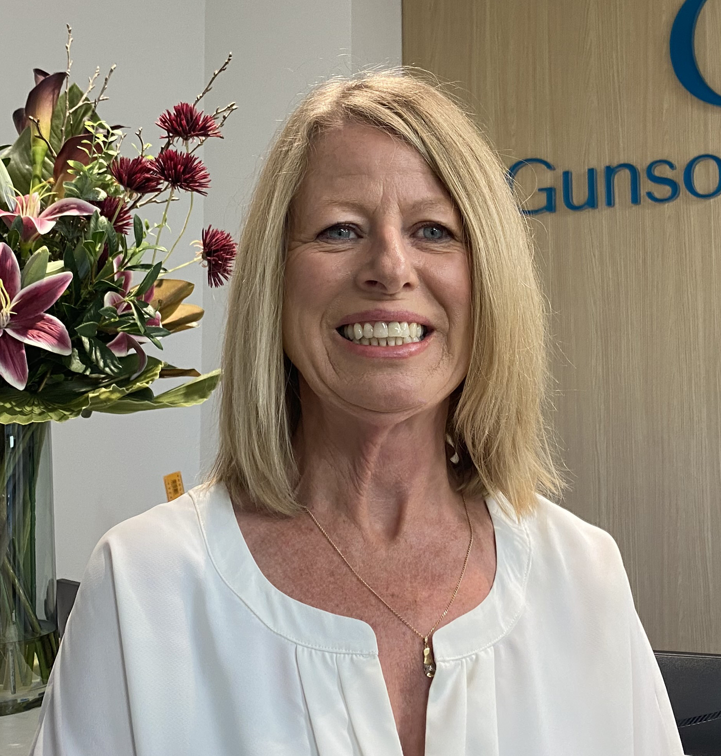 Woman smiling, wearing a white shirt, with blonde hair, in an office setting with flowers.