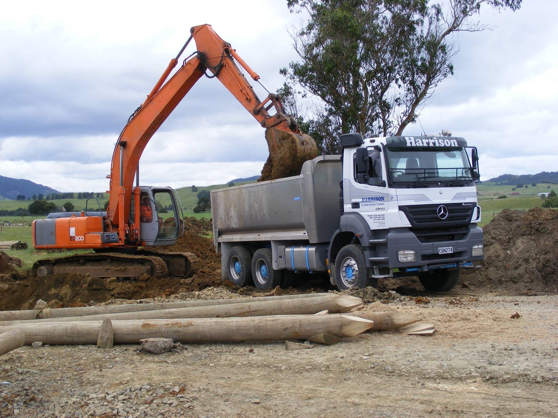 An excavator loading dirt into a dump truck in a rural setting under a cloudy sky.