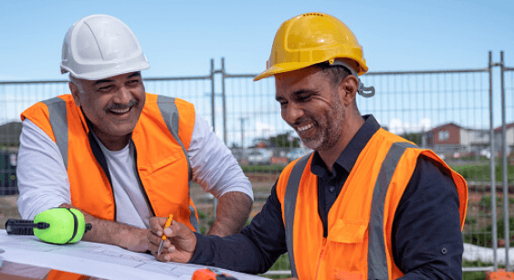 Two construction workers in vests and helmets laughing over blueprints outdoors.