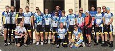 A group of cyclists wearing matching blue and white jerseys, some with medals, posing outside in front of a building.