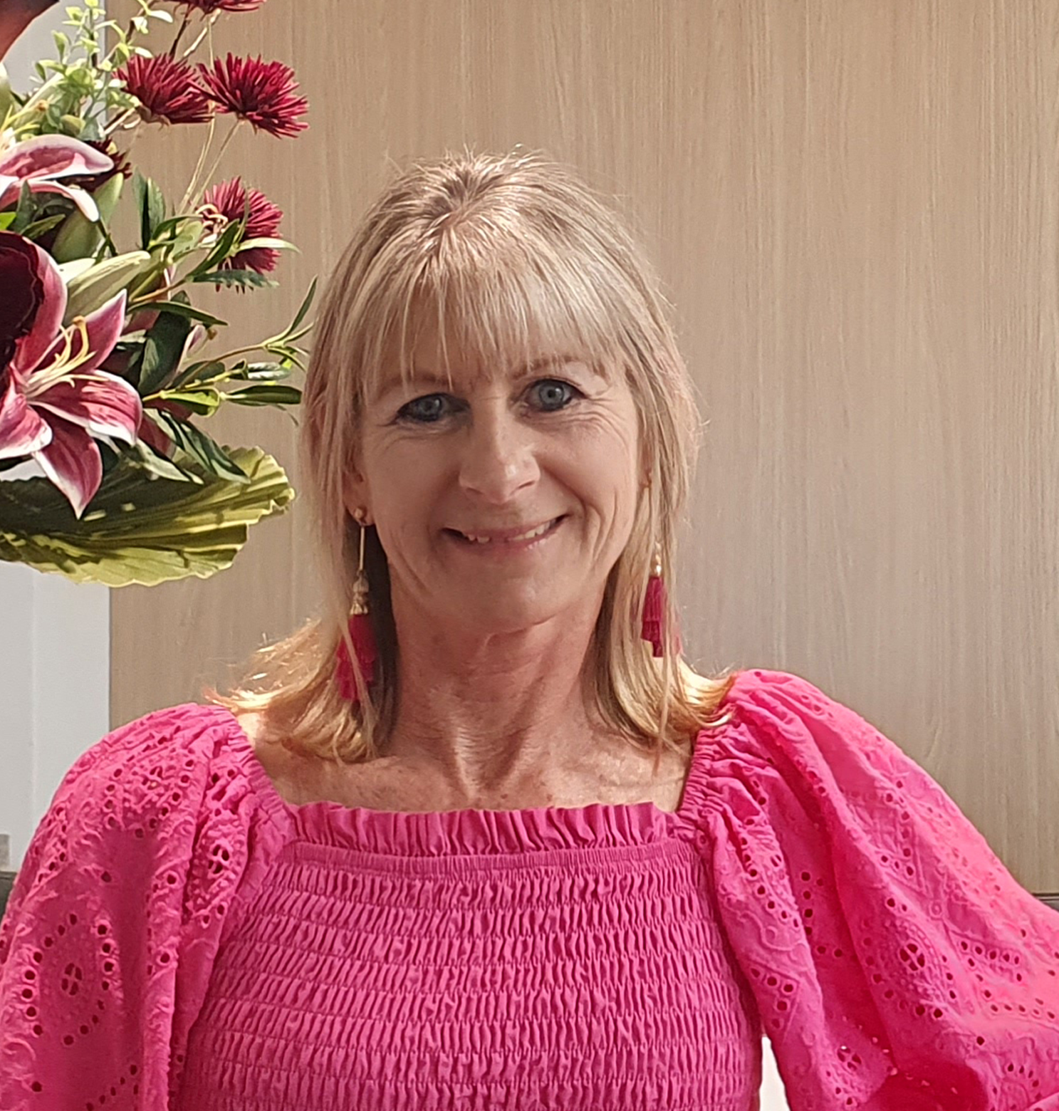 Woman with blonde hair, smiling in a pink eyelet top, near floral arrangement.