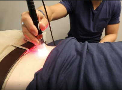 A person using a handheld device on a patient's knee. The device has a red light. Indoors.