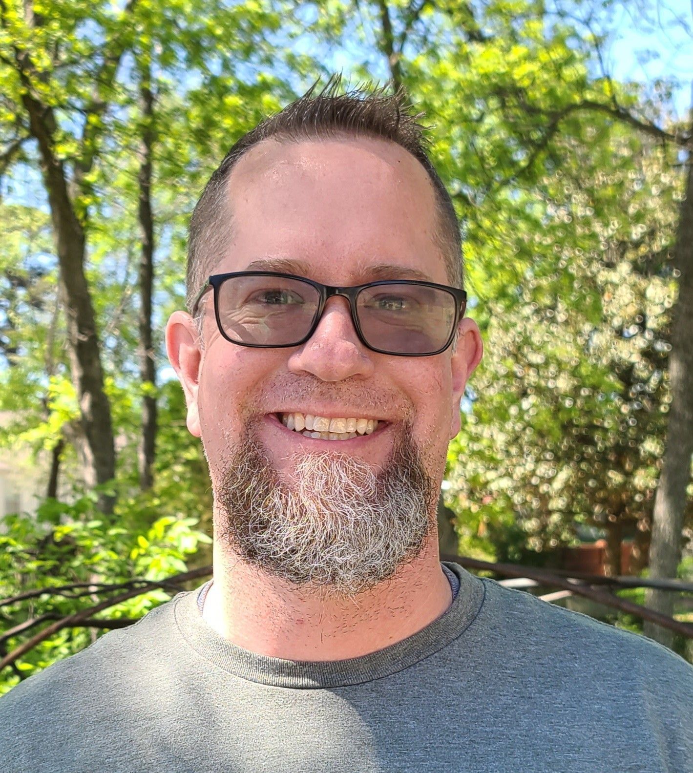 Smiling man with glasses and a gray shirt outdoors in a sunny, leafy setting