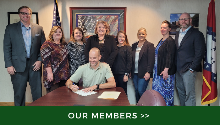 Group photo of eight people in an office, with one man seated at a desk signing papers, and a “Our Members” banner below
