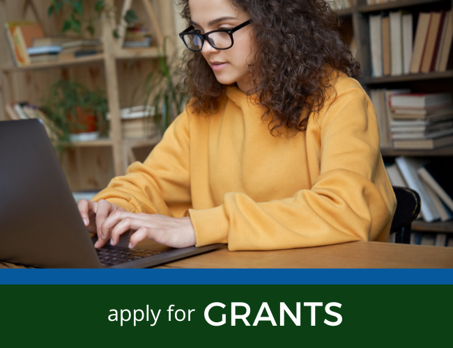 Woman in yellow sweater typing on a laptop with “apply for GRANTS” banner below