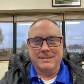 Person wearing glasses and a blue shirt, seated in an office with windows behind them.