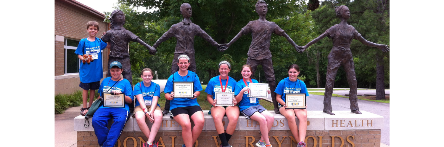Team in blue shirts posing on a bench before a bronze statue with outstretched arms.