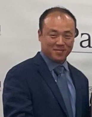 Smiling man in a navy suit and blue tie standing before a white backdrop with partial text